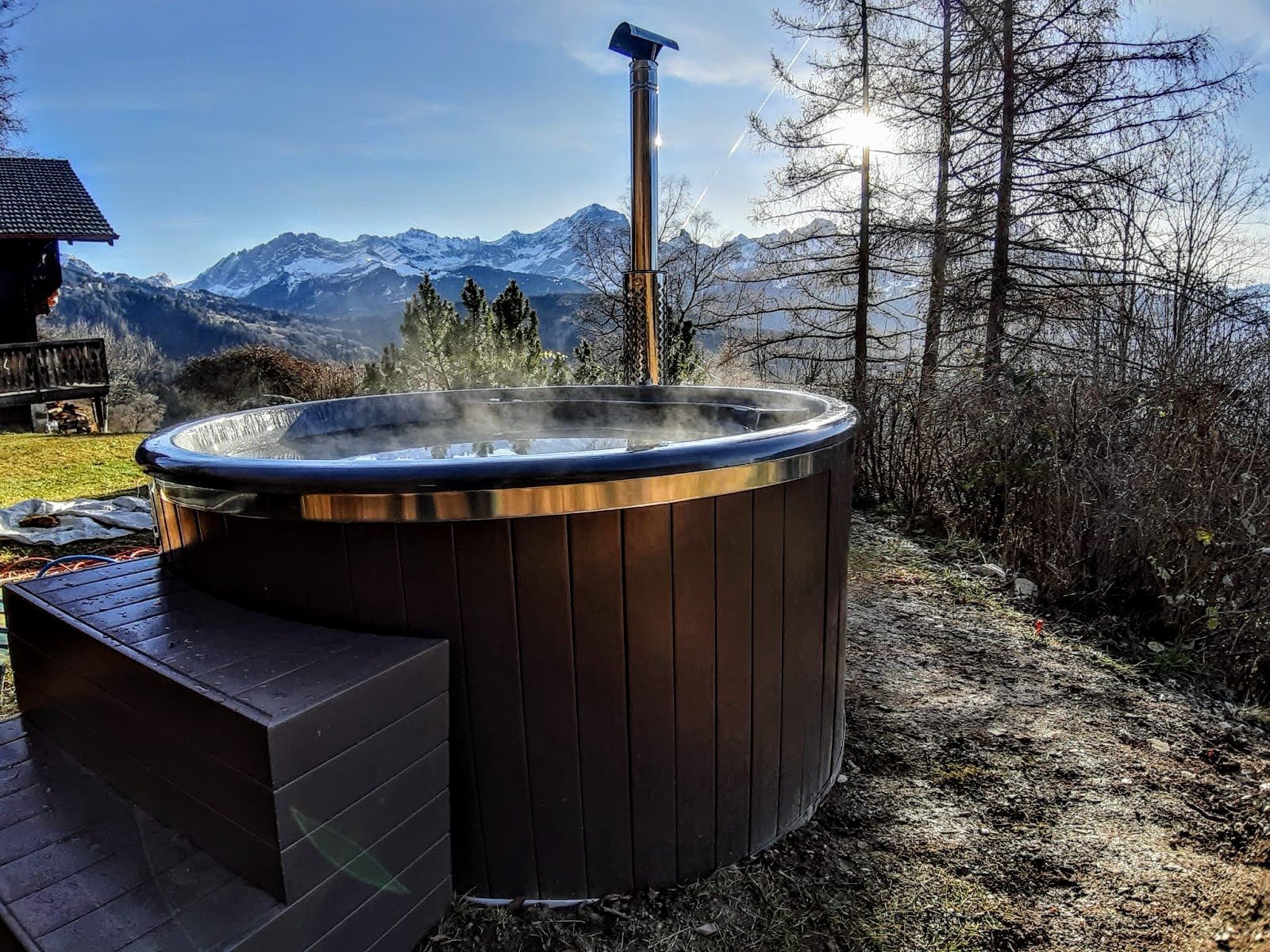 TimberIN UK 1 Steaming round wood-fired hot tub with external stove and stainless chimney in front of snow-capped mountains