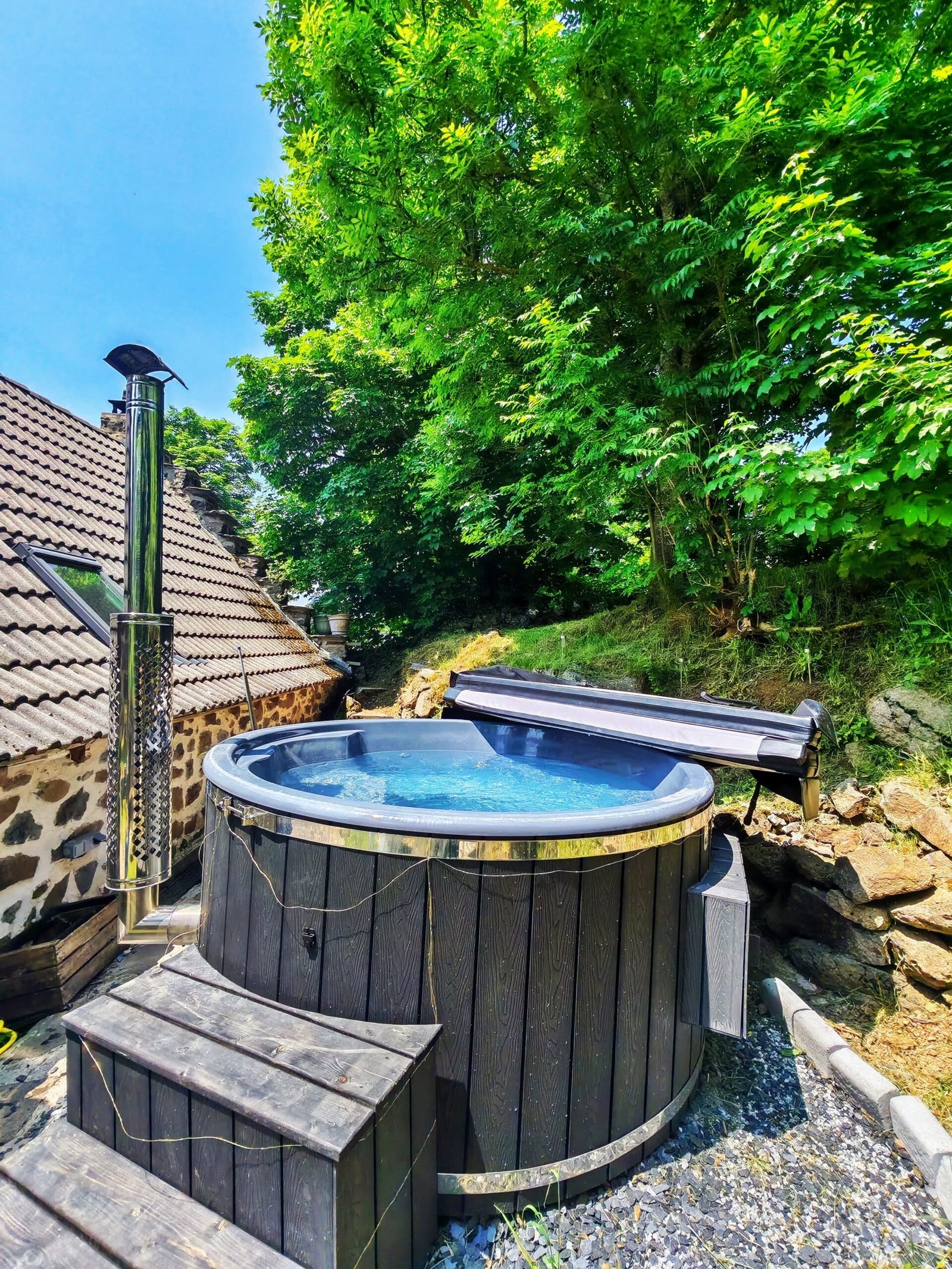 Wood Fired Heater for Hot Tub 1 Dark timber-clad hot tub with a chimney beside a stone building and trees