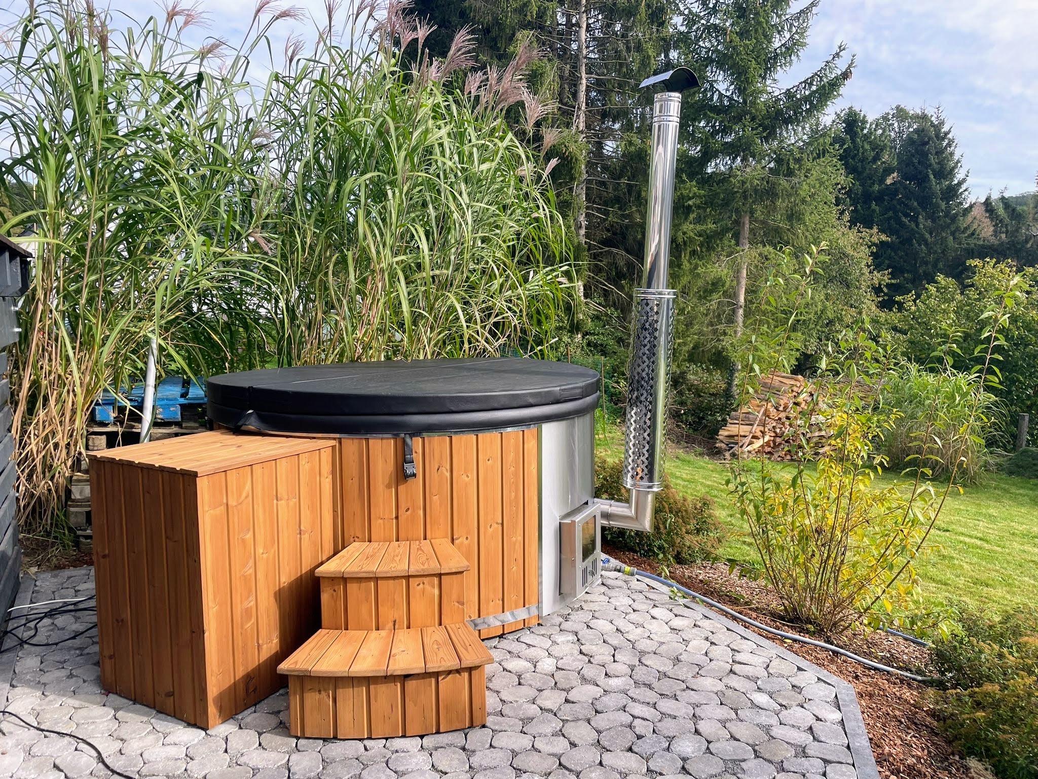 Round timber-clad hot tub with a black cover, wooden steps and a stainless-steel chimney on a paved garden base