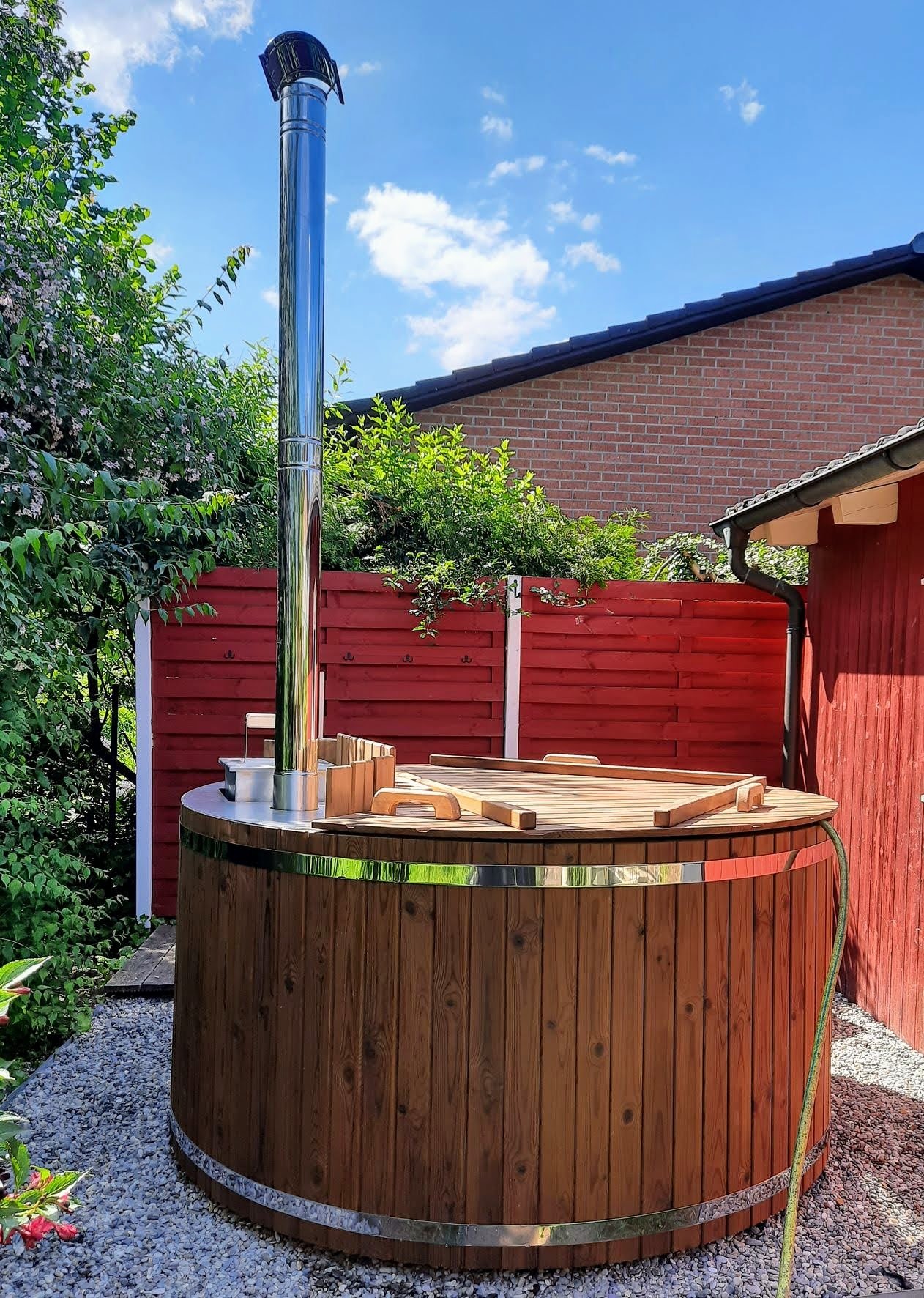 Wood Fired Soaking Tubs 1 Round wooden hot tub with stainless-steel bands, a wooden lid and tall chimney beside a red garden fence