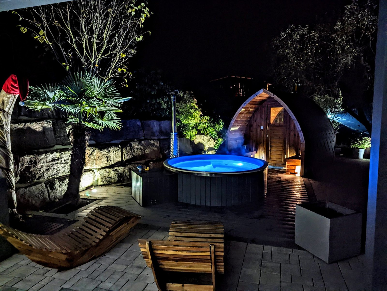 Sauna Hot Tub Combo 1 Blue-lit round hot tub beside a wooden pod sauna, with garden seating, timber decking and a planted stone boundary at night
