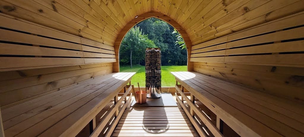 Electric Outdoor Saunas 1 Timber sauna interior with benches on both sides, a cylindrical stone heater and an arched window overlooking a green garden