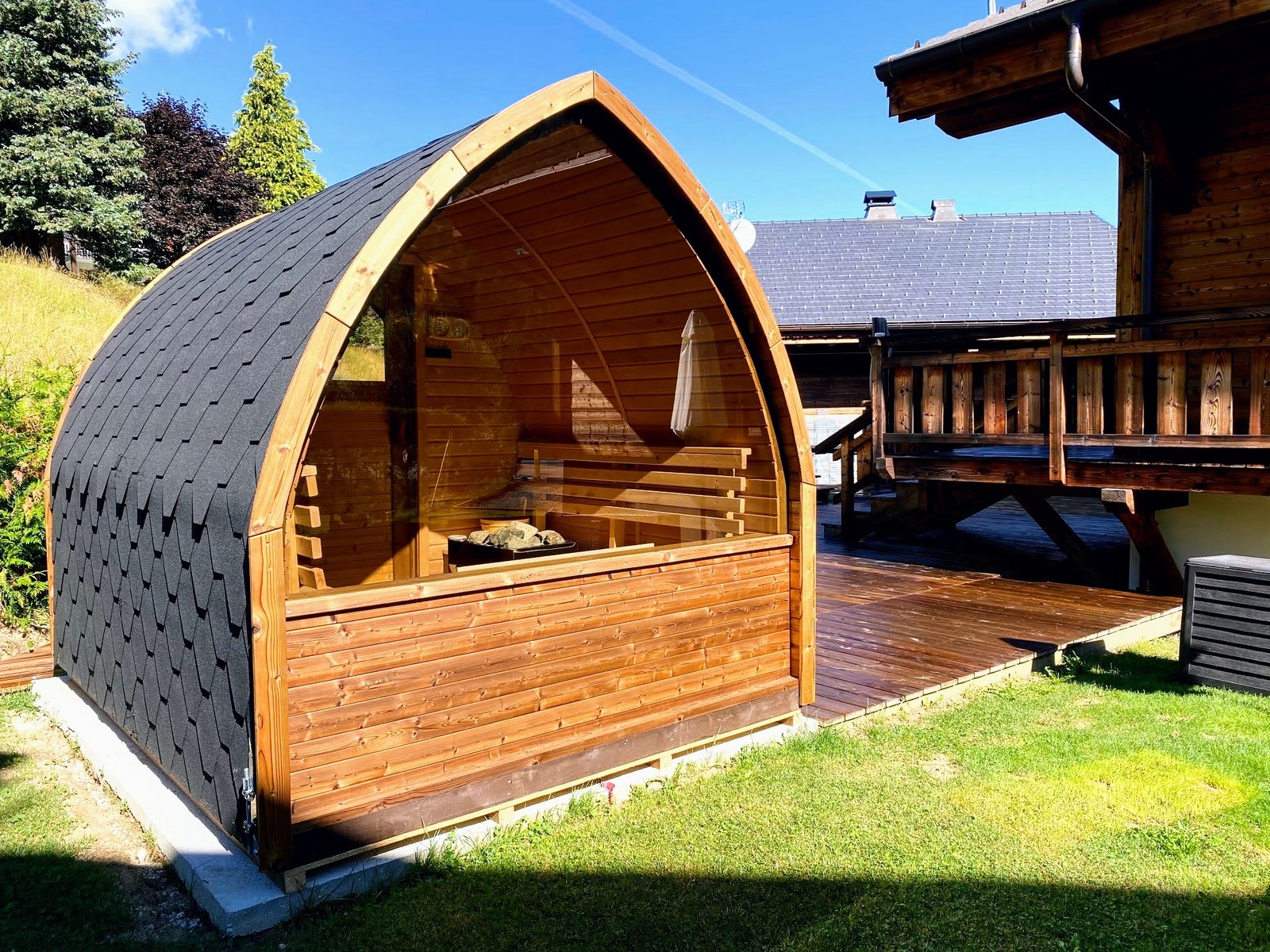 Prices 1 Timber sauna with a glazed front, visible bench and stone heater, shingled roof and base beside wooden decking