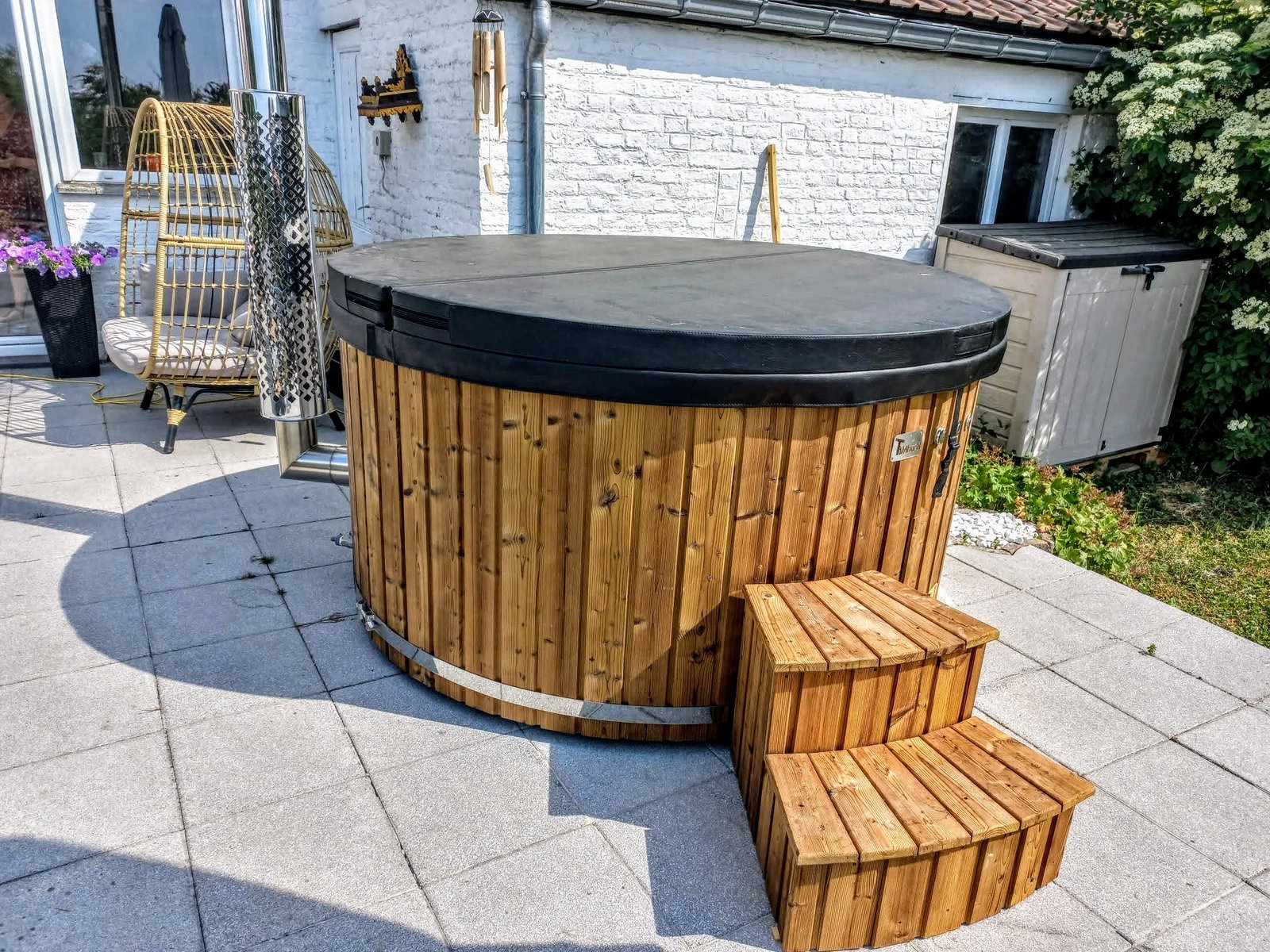 Affordable Hot Tubs UK: How to Buy Value, Not a False Economy 1 Round timber-clad hot tub with a closed dark cover, wooden access steps and a shielded metal flue on a paved patio
