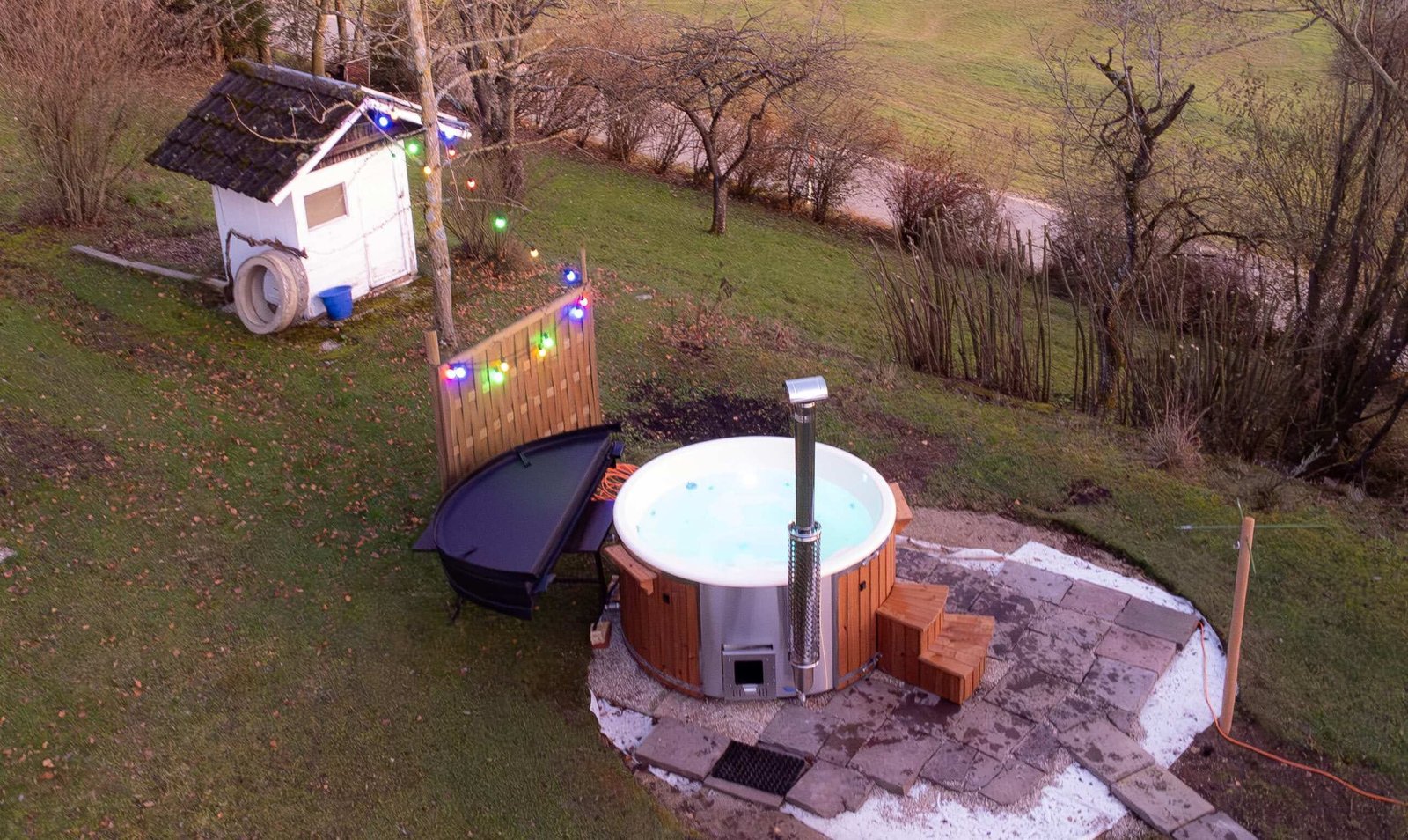 Round Hot Tubs 1 Elevated view of a round timber-clad hot tub with a pale liner, access steps, metal flue and folded cover beside it