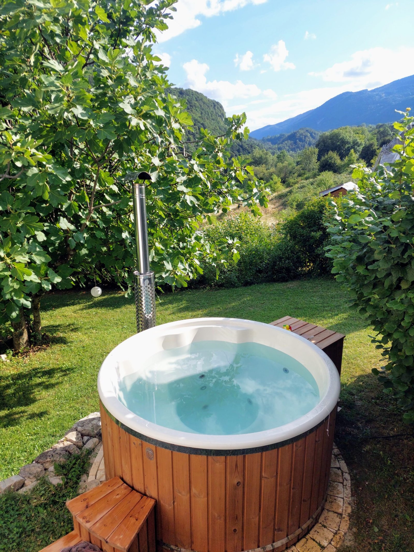 8 Person Hot Tubs UK: Large-Capacity Project Guide 1 Round timber-clad hot tub with a white moulded interior, water, wooden entry steps and a metal flue in a planted garden