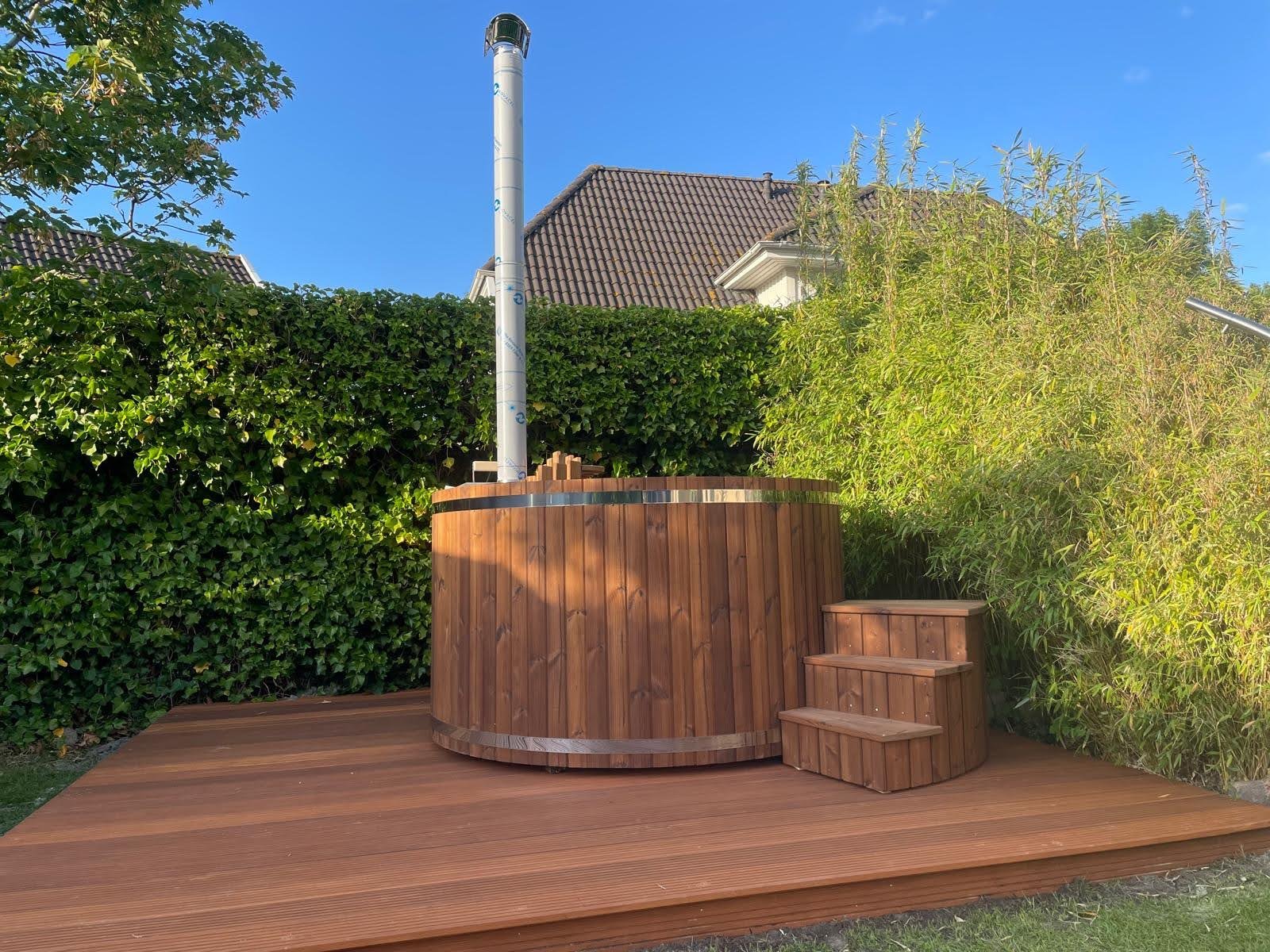 Garden Hot Tub 1 Round timber hot tub with wooden entry steps and a tall metal flue on decking beside a hedge and bamboo