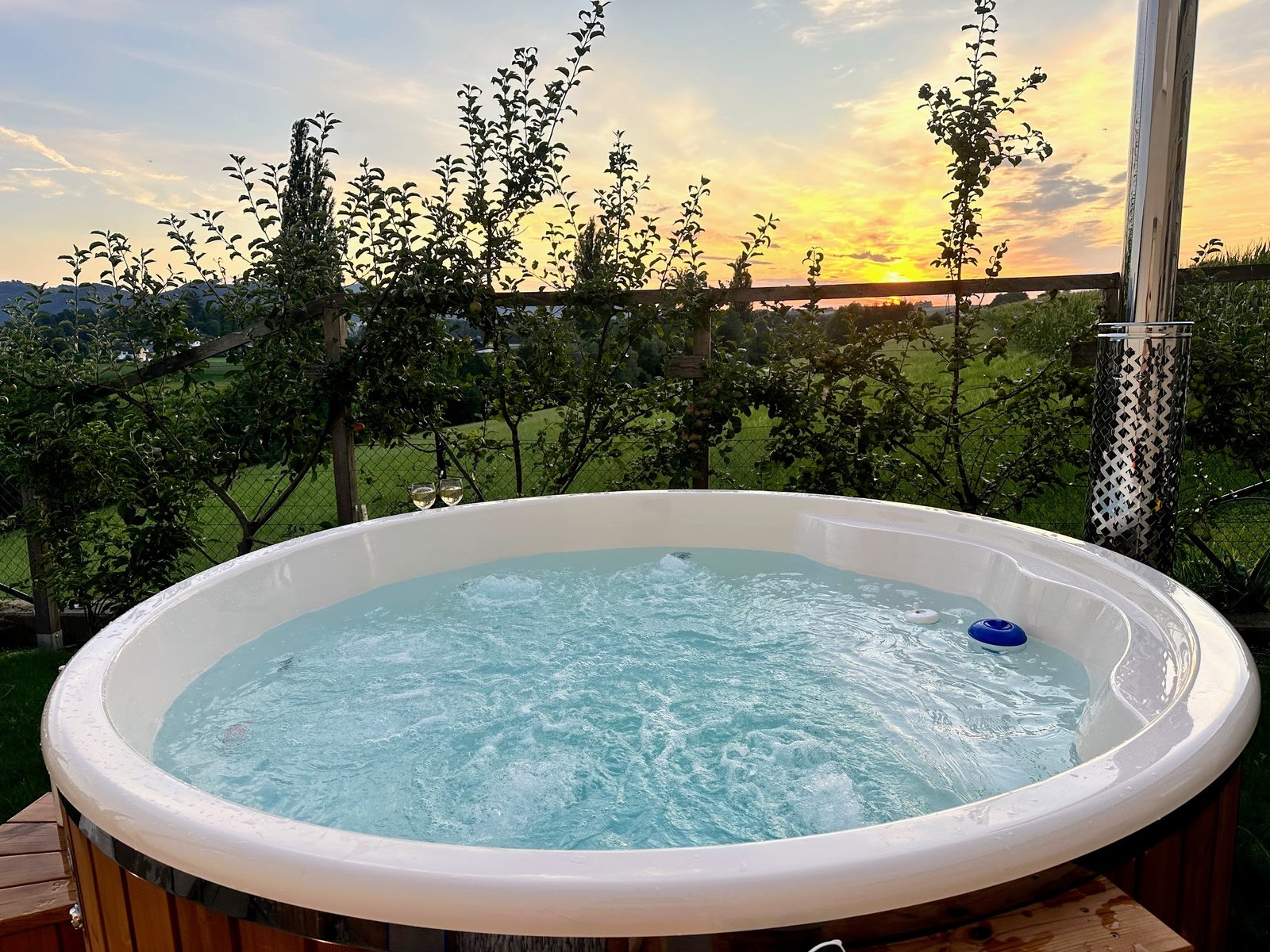 Round hot tub with a white liner, moving water and a guarded metal flue beside garden planting at sunset