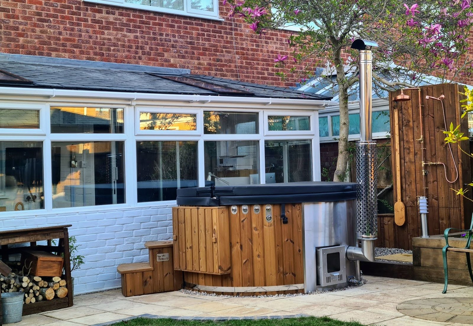 Copper-coloured shower head and handset on timber screening beside a covered round hot tub, paved patio and flowering tree