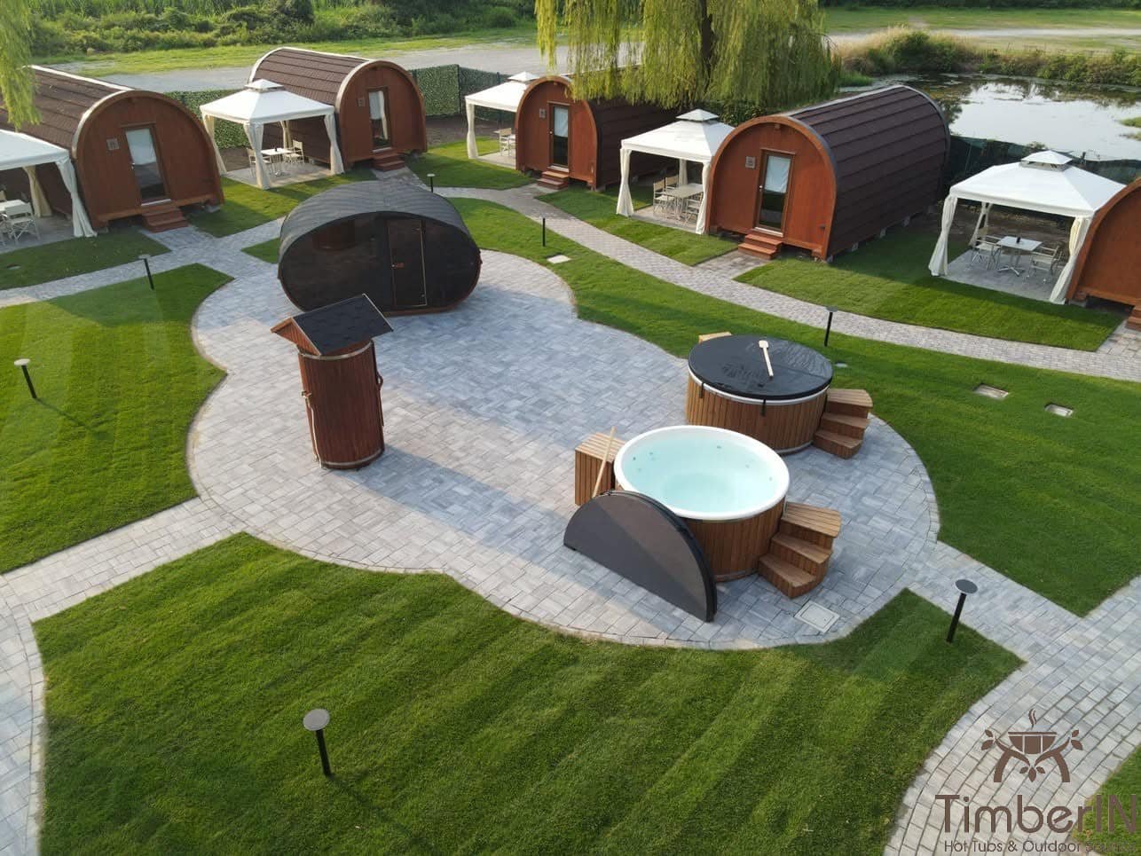 Aerial view of curved timber buildings, lawns and paths around a paved display area with hot tubs and an outdoor shower