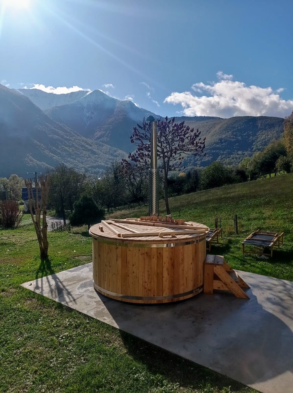 Round wooden hot tub with a timber lid, steps and metal chimney on a level platform overlooking mountains