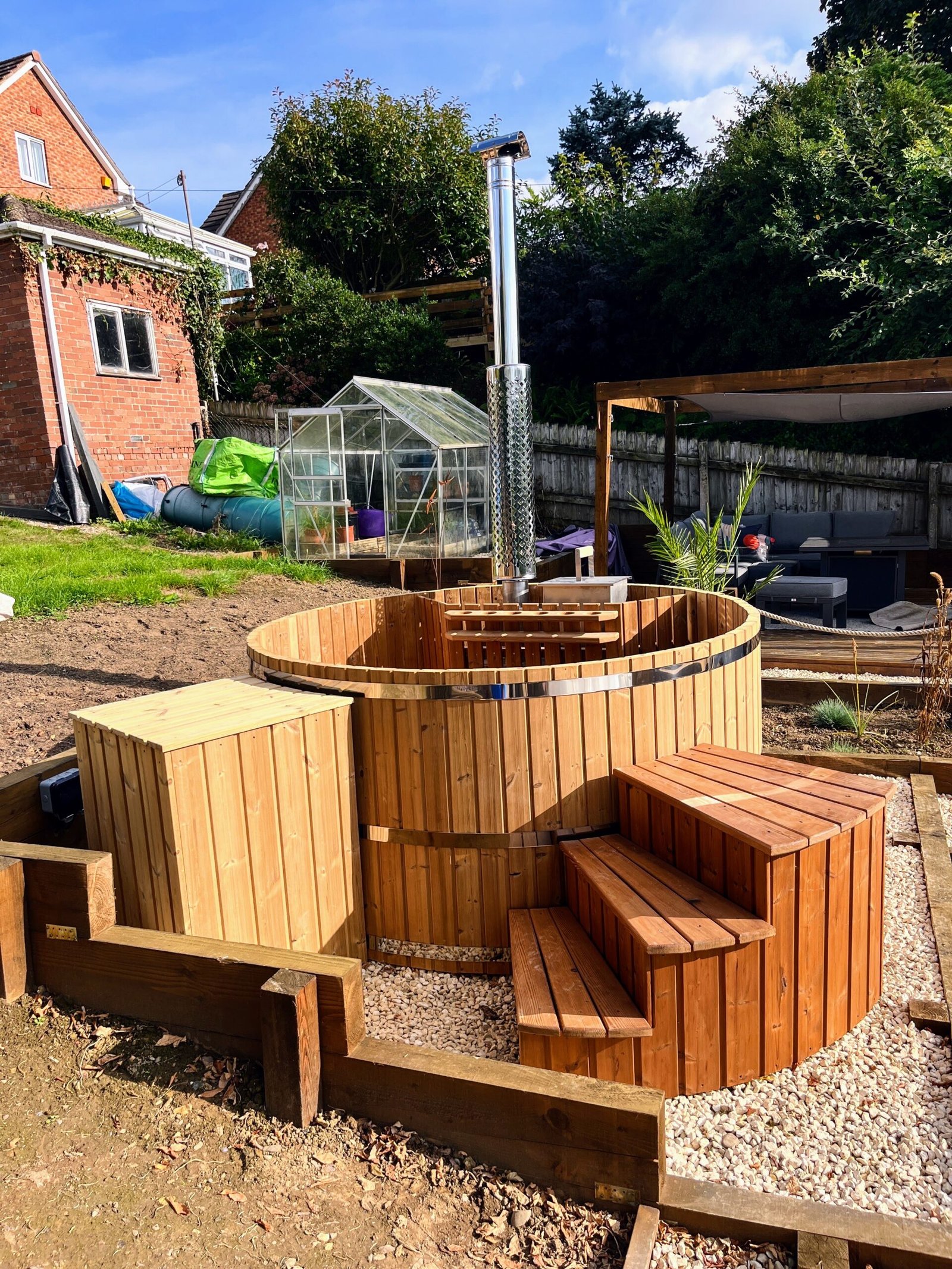 salty-barrel-hot-tubs 1 Round wooden hot tub with an internal stove guard, tall metal chimney and broad wooden steps in a landscaped garden