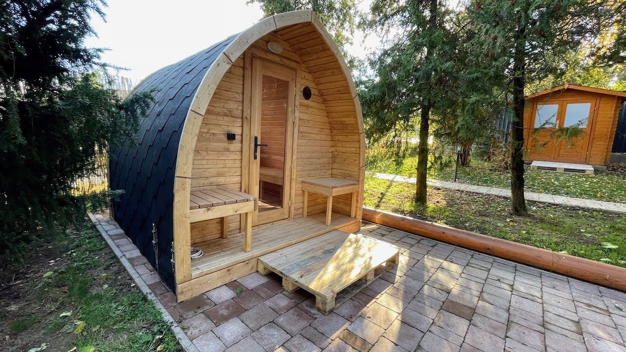 Pod 1 Timber sauna pod with a dark curved roof, glazed entrance door, two porch benches and a wooden step