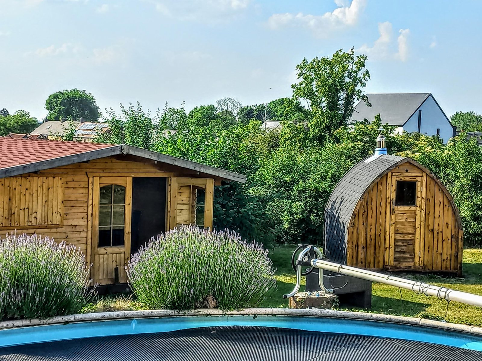 Best Outdoor Sauna 1 Curved-roof timber sauna beside another garden building, shrubs and the edge of a covered pool