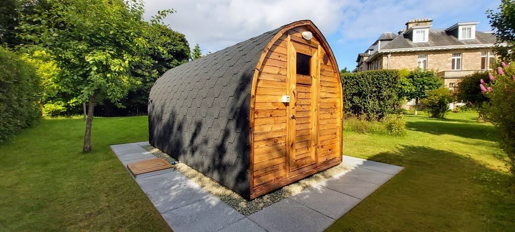 Near Me 1 Timber pod sauna with a dark roof on a gravel base surrounded by paving in a landscaped garden