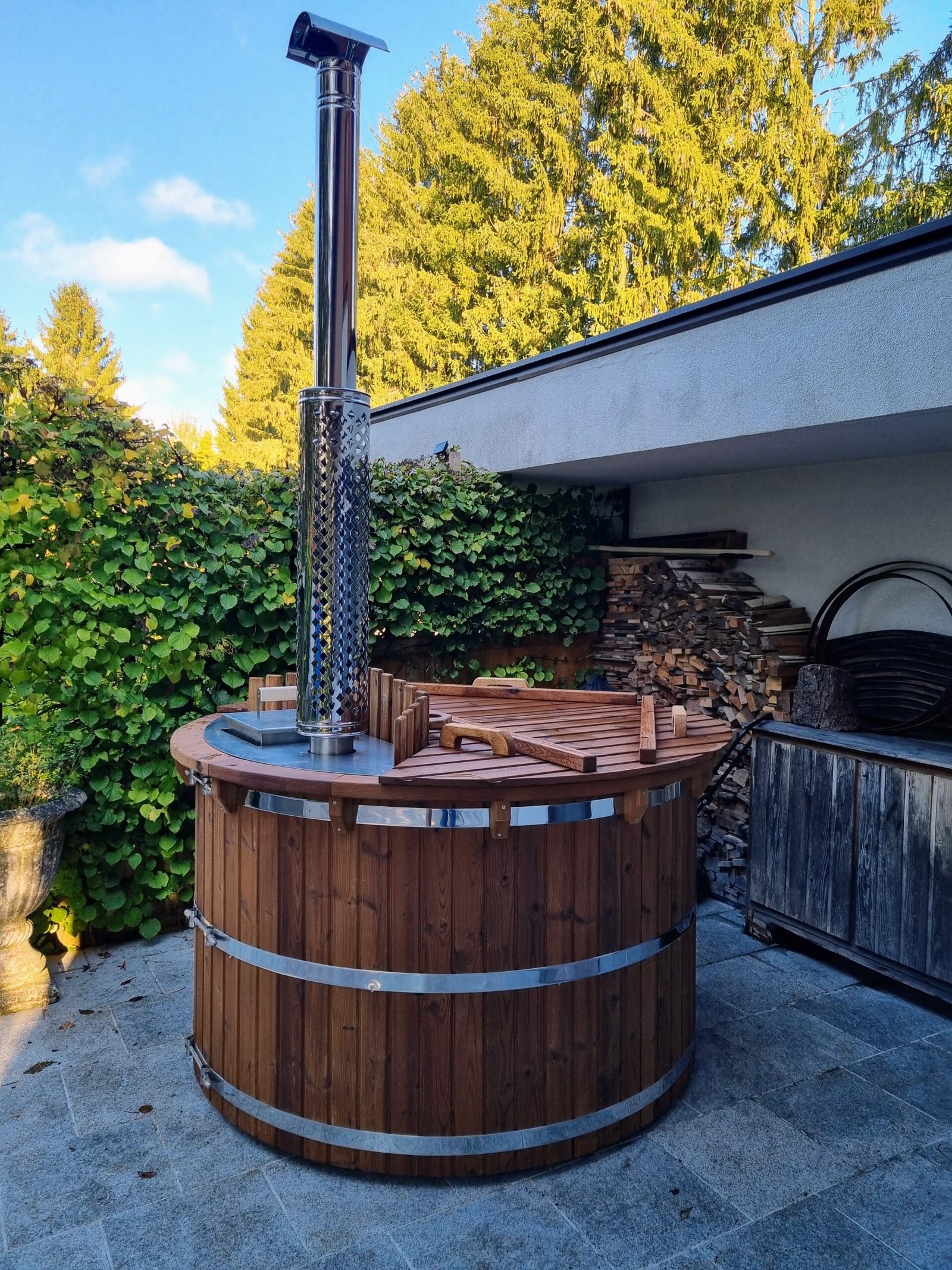 Full view of a round TimberIN wooden hot tub with timber lid, internal wood-fired heater, complete chimney and garden setting